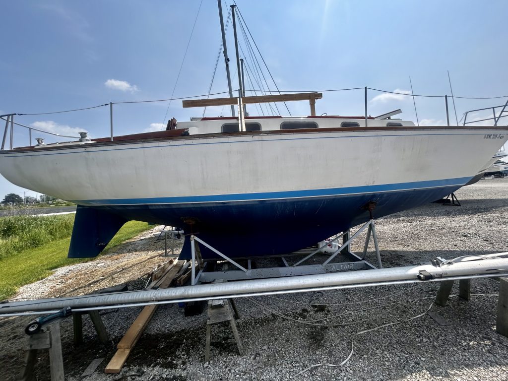 A dirty old sailboat, mast horizontal in front of the hull, on its cradle in a gravel lot