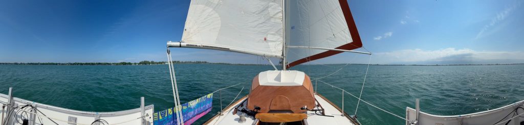 A panoramic view of Altair, wing in wing, sailing north in the Detroit River.  Solar panels and a towel are visible on the life lines, and our mahogany dinghy occupies the center of the deck.